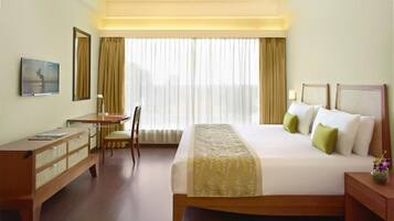 Una habitación de hotel con cama, escritorio, silla y un amplio ventanal con cortinas.