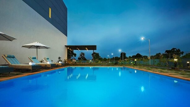 A swimming pool with lounge chairs and umbrellas, surrounded by a building with a flat roof. - Ginger Gondia, Balaghat Road (Gondia)