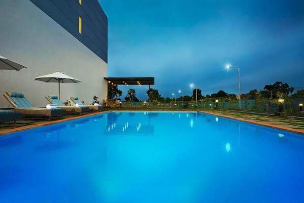 A swimming pool with lounge chairs and umbrellas, surrounded by a building with a flat roof. - Ginger Gondia, Balaghat Road (Gondia)