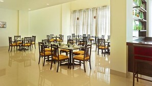 A spacious dining area with multiple tables and chairs, a large window with curtains, and a shelf with green bottles. - Ginger Gondia, Balaghat Road (Gondia)