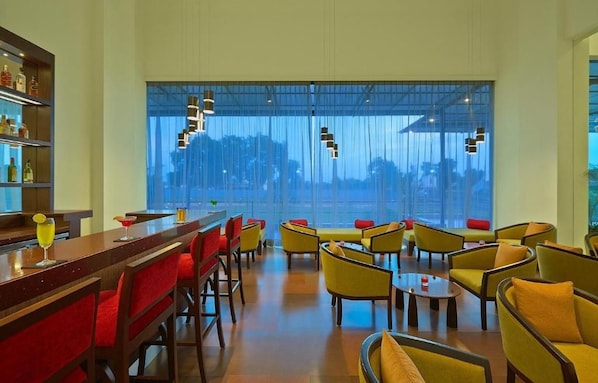 A bar area with a long counter and red upholstered chairs, a seating area with yellow chairs and tables, and a large window with a view of trees and buildings. - Ginger Gondia, Balaghat Road (Gondia)