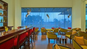 A bar area with a long counter and red upholstered chairs, a seating area with yellow chairs and tables, and a large window with a view of trees and buildings. - Ginger Gondia, Balaghat Road (Gondia)