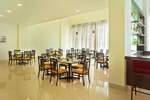 A spacious dining area with multiple tables and chairs, a large window with curtains, and a shelf with green bottles. - Ginger Gondia, Balaghat Road (Gondia)