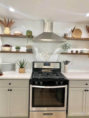 A kitchen with a stainless steel stove, oven, and a modern range hood. There are wooden shelves above the stove with various kitchen items and plants.