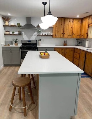 Een keuken met een centraal eiland, houten kastjes, een fornuis en een hangende lamp.