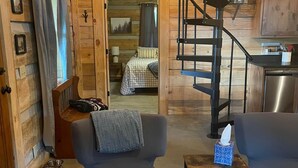 A cozy cabin interior with a modern armchair, a small table with a tissue box, a pet bowl, and a spiral staircase leading to a bedroom. - Cozy, Quiet Cabin Under the Stars - Near Desoto State Park and Downtown Mentone! (Mentone)