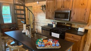 A kitchen with wooden cabinets, a microwave, a stove, a sink, and a bar area with chairs. - Cozy, Quiet Cabin Under the Stars - Near Desoto State Park and Downtown Mentone! (Mentone)