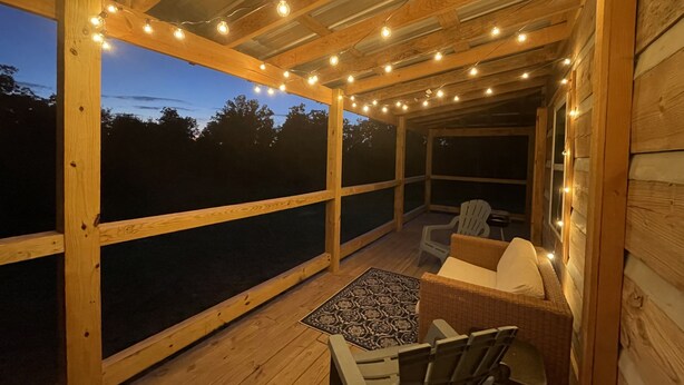 A wooden deck with string lights, a wicker sofa, a table, and a view of the outdoors. - Cozy, Quiet Cabin Under the Stars - Near Desoto State Park and Downtown Mentone! (Mentone)
