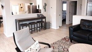 Un salon moderne avec un canapé en cuir noir, un fauteuil blanc et un tapis à motifs. On aperçoit à l’arrière-plan un coin cuisine avec un bar et des tabourets.