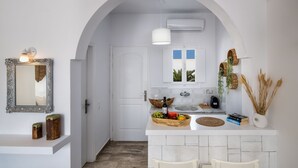 A kitchen with white cabinetry, a white countertop, and a white archway. There is a mirror with a decorative frame, a basket of fruit, and a vase with dried plants. - Sol Mare Salis (Santorini)