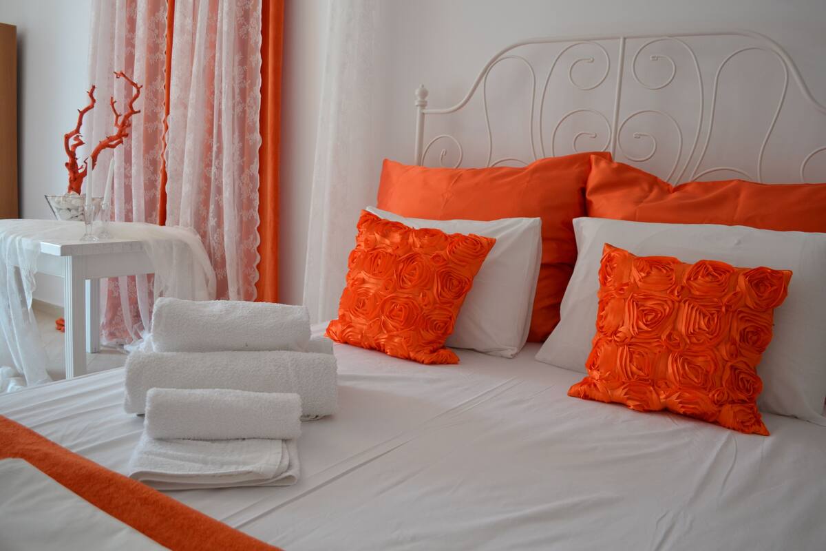 A neatly made bed with white and orange bedding, a white headboard, and a small table with a vase and towels.