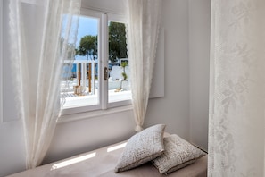 Una habitación con ventana, cortinas blancas, una cama con almohadas estampadas y vista al exterior.