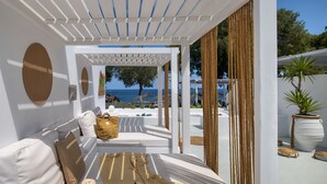 A sunlit outdoor seating area with a white sofa, a woven basket, and a book. A view of the sea and beach is visible through the pergola. - Sol Mare Salis (Santorini)