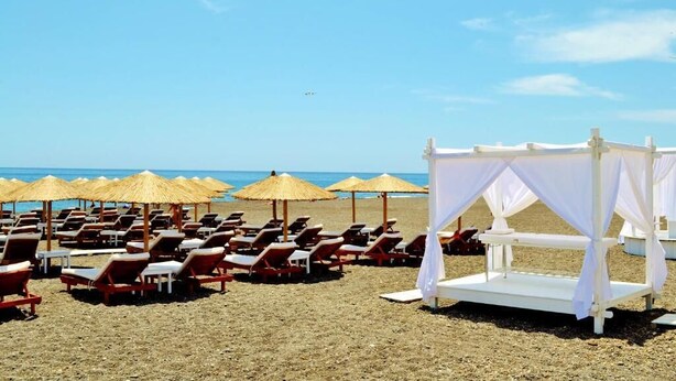 A beach with rows of sun loungers and thatched umbrellas, a white canopy structure, and the ocean in the background. - Sol Mare Salis (Santorini)