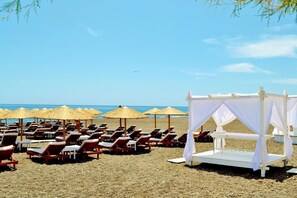 A beach with rows of sun loungers and thatched umbrellas, a white canopy structure, and the ocean in the background. - Sol Mare Salis (Santorini)
