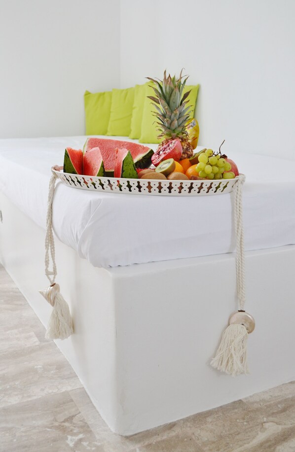 A bed with a white mattress, a decorative tray with fruits and a pineapple, and a green pillow. - Sol Mare Salis (Santorini)