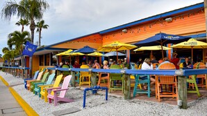A beachside restaurant with outdoor seating, palm trees, and colorful chairs.