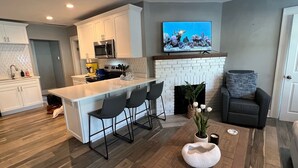 A modern kitchen with white cabinets, a central island with bar stools, a flat-screen TV above the fireplace, and a cozy seating area with a couch and a decorative plant.