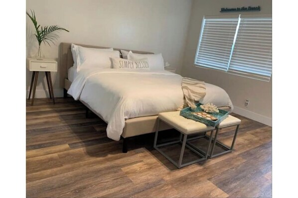 Quarto com cama, mesa de cabeceira, banqueta e um letreiro na parede.