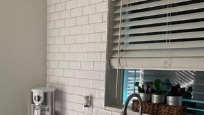 Una cocina con baldosas blancas estilo metro, un lavamanos de acero inoxidable, una cafetera y un ventanal con persianas.