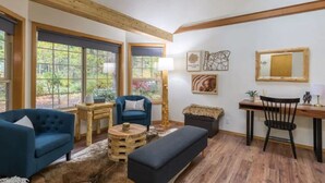 Ein gemütliches Wohnzimmer mit Holzboden, großen Fenstern, einem blauen Sofa, einem runden Couchtisch und einem Schreibtisch mit Stuhl.