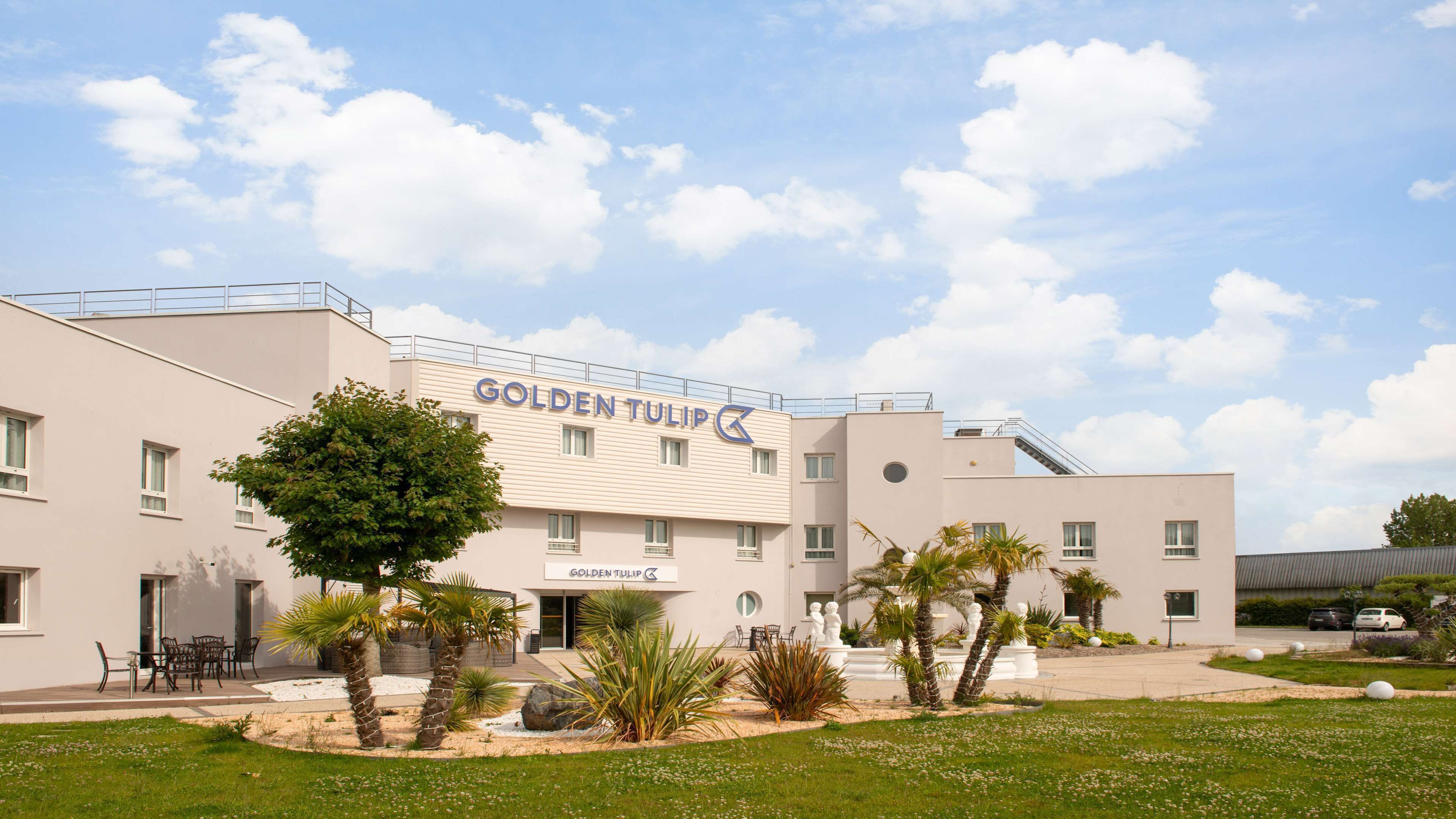 A hotel named Golden Tulip with a modern architectural design, featuring a spacious outdoor area with palm trees and a well-maintained garden.
