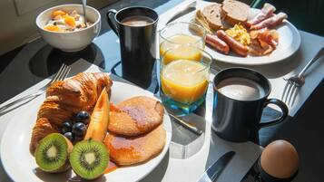 Une table de petit-déjeuner avec des croissants, des kiwis, des myrtilles, des crêpes, du jus d’orange, du café et une assiette d’œufs, de bacon et de pain grillé.