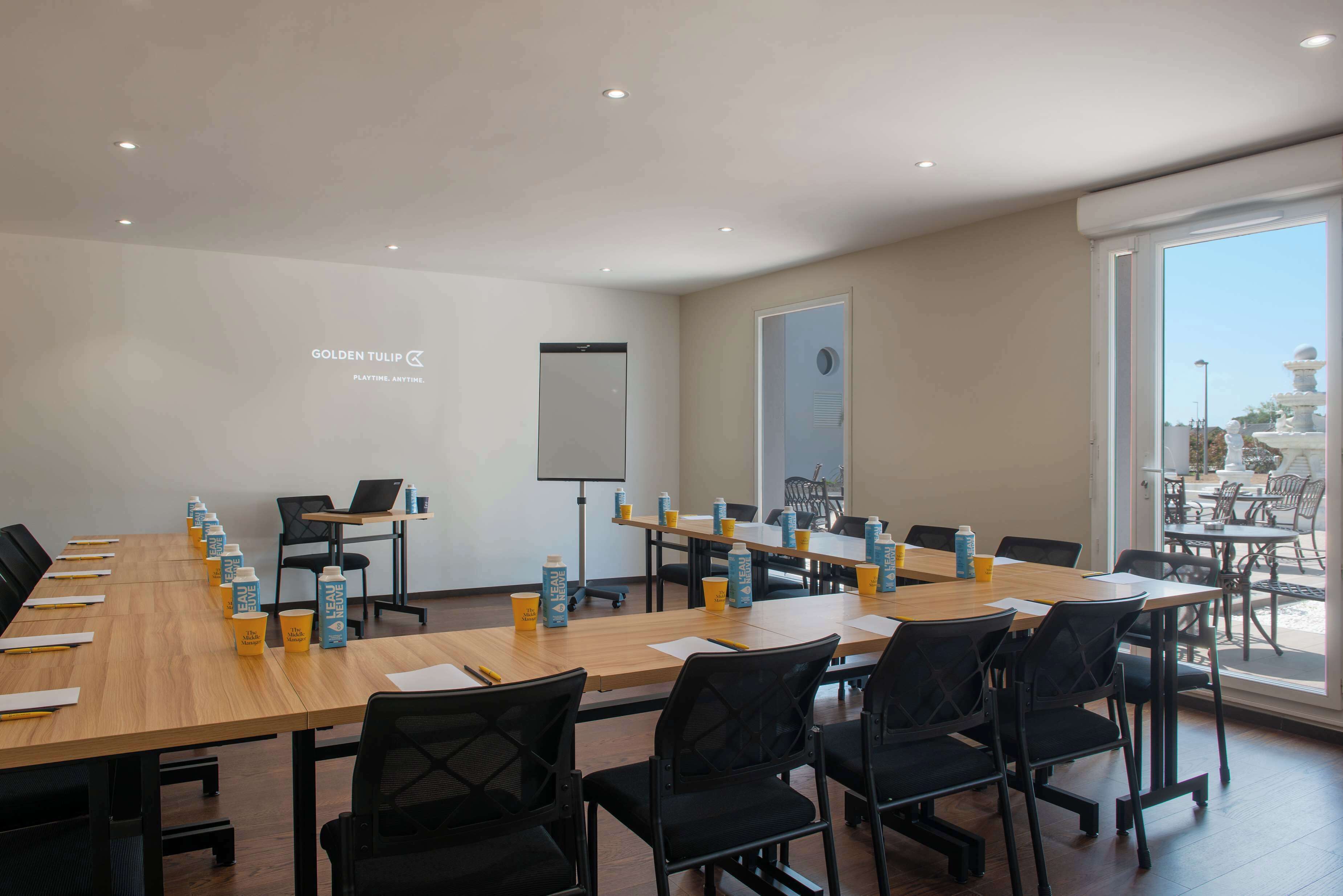 Une salle de conférence équipée d’une longue table, de chaises, d’un écran de projection et d’une grande fenêtre donnant sur un espace extérieur aménagé avec des sièges.