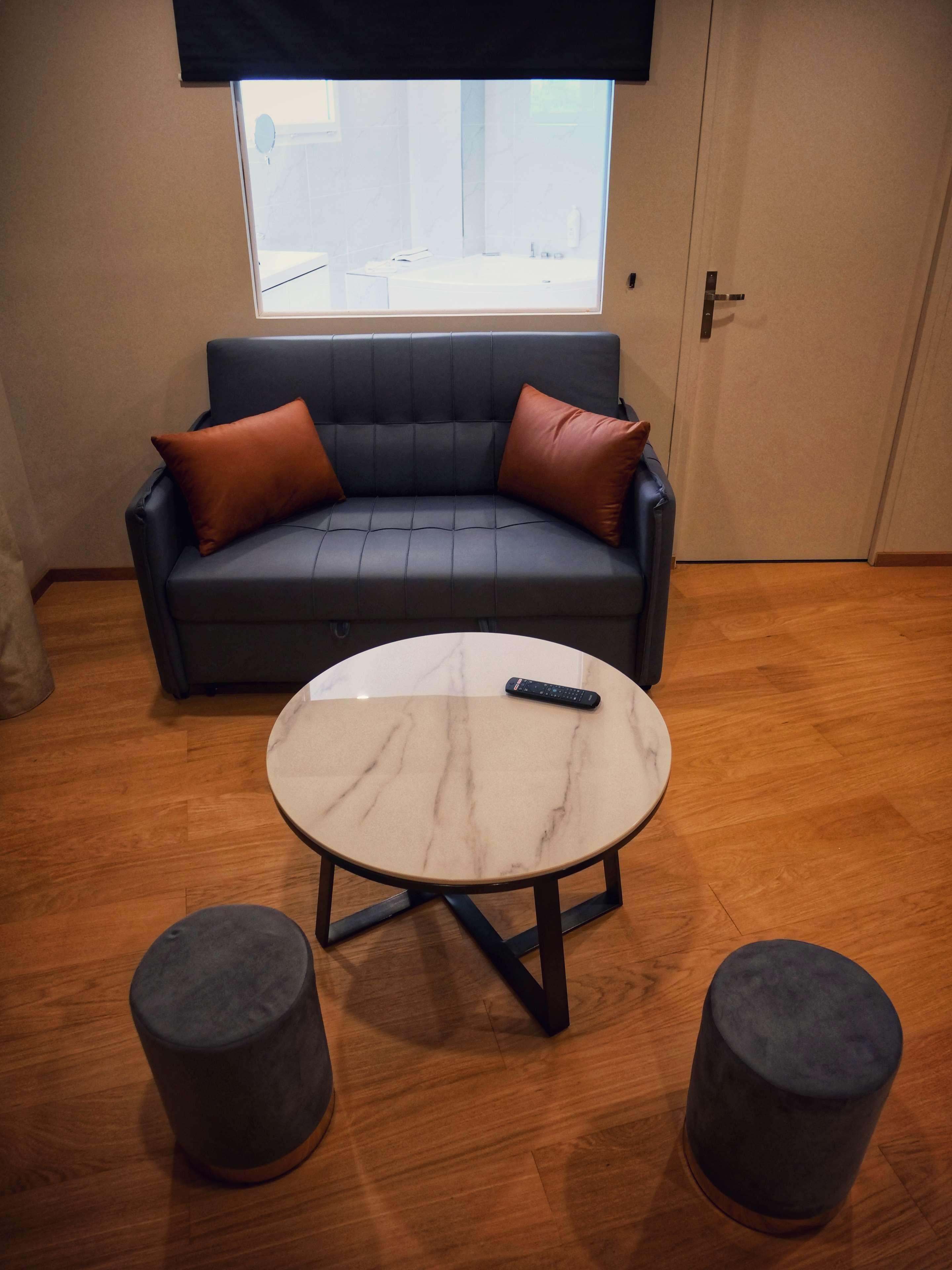 A room with a dark blue sofa with brown cushions, a round marble table with two grey stools, and a window above the sofa.