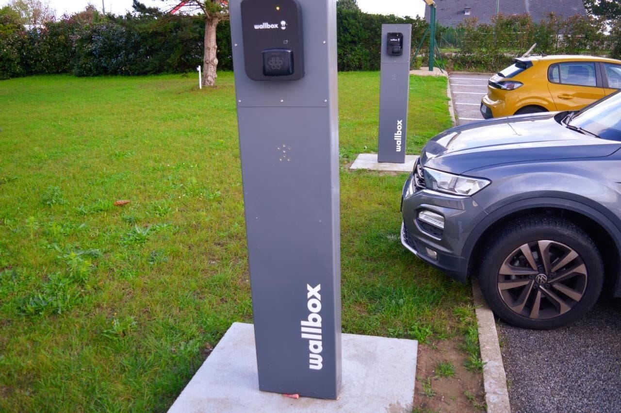 A gray wallbox with the 'wallbox' logo, a parking area with a yellow car, and a gray SUV.