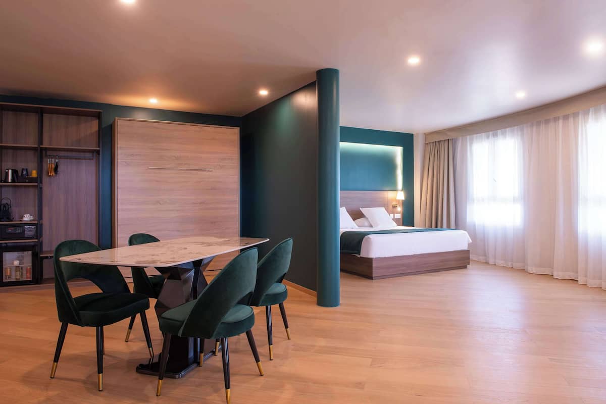 A modern hotel room with a dining area, a bed with a headboard, and a large window with curtains.