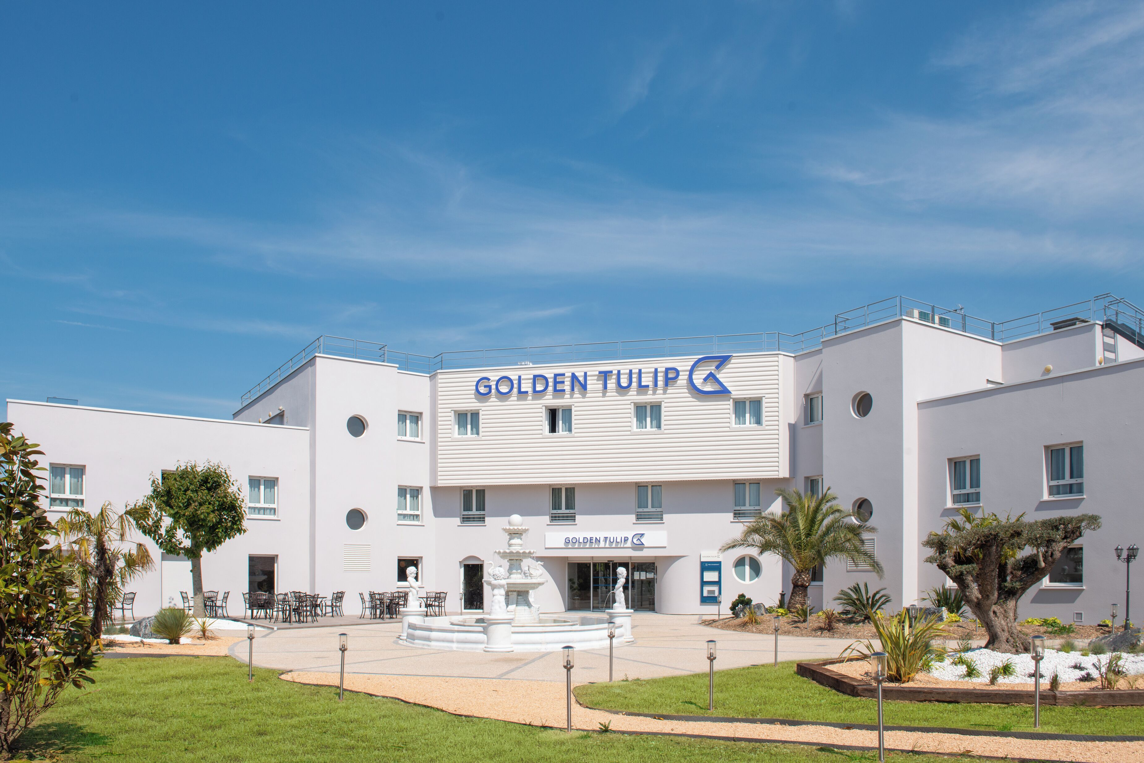 Un hotel moderno llamado "Golden Tulip" con una fuente central, rodeado de jardines bien cuidados y palmeras.