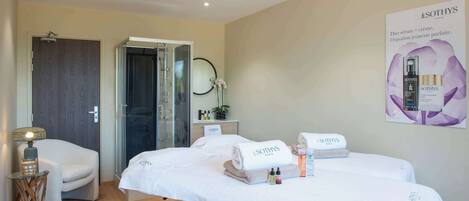 A spa room with two beds, a massage table, a small table with a lamp, and a poster on the wall.
