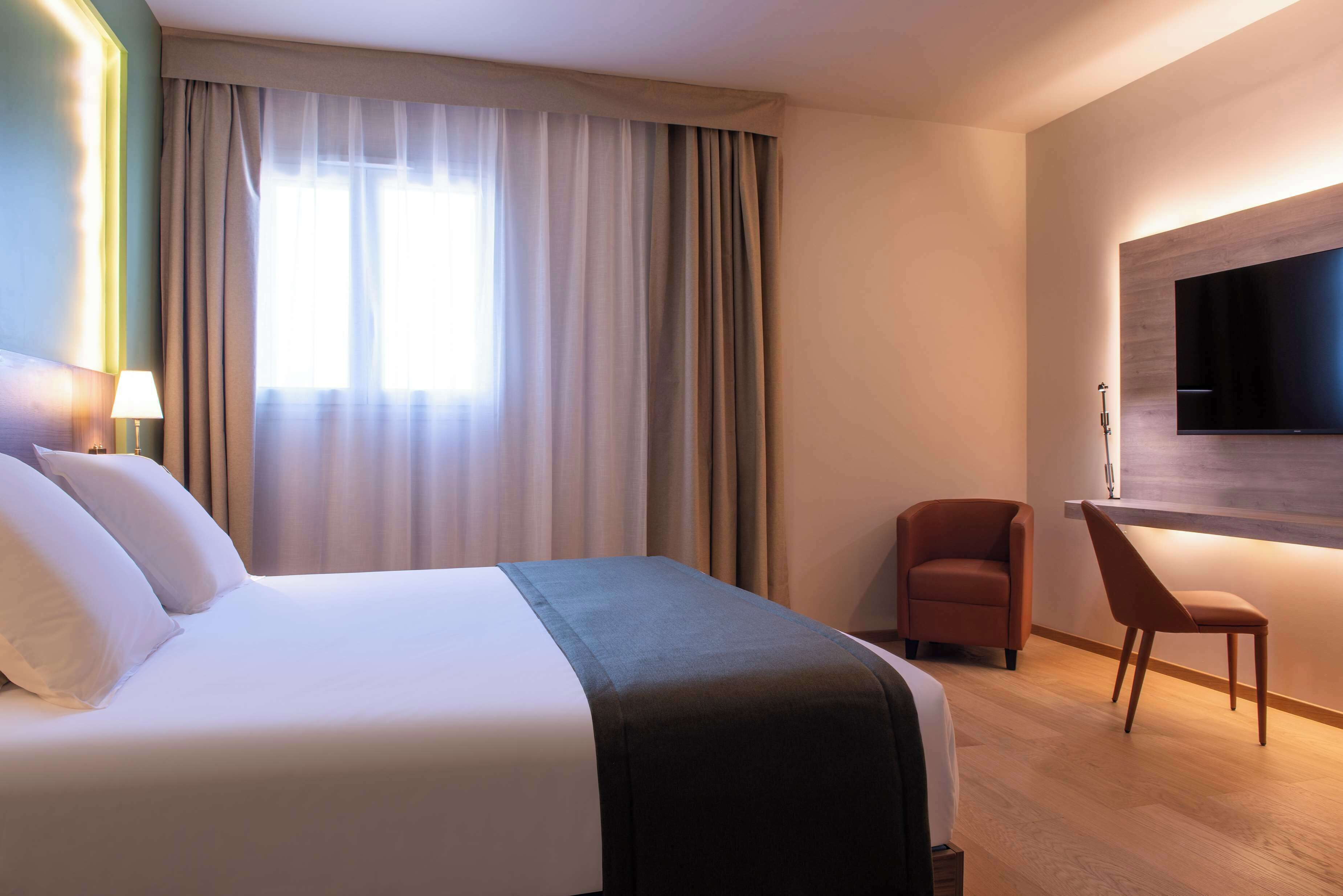 A hotel room with a large bed, a desk, a chair, and a flat-screen TV mounted on the wall.