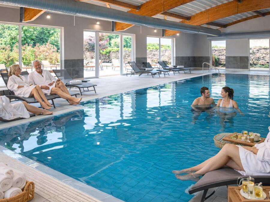 An indoor swimming pool with lounge chairs, a sunbed, and a poolside area with a view of greenery.