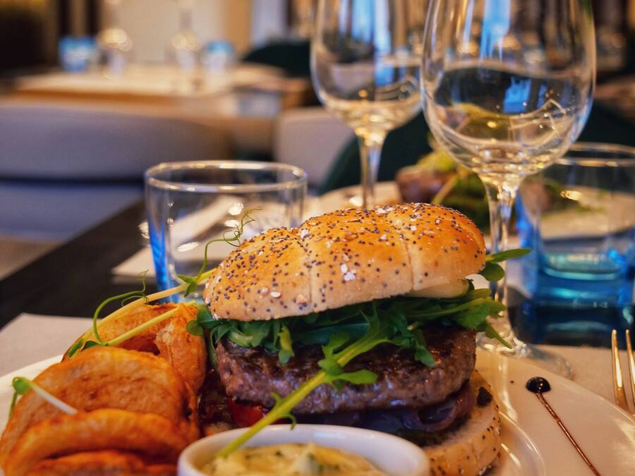 Una mesa de comedor con una hamburguesa, papas fritas y un recipiente pequeño de salsa. Hay copas de vino y una botella de agua sobre la mesa.