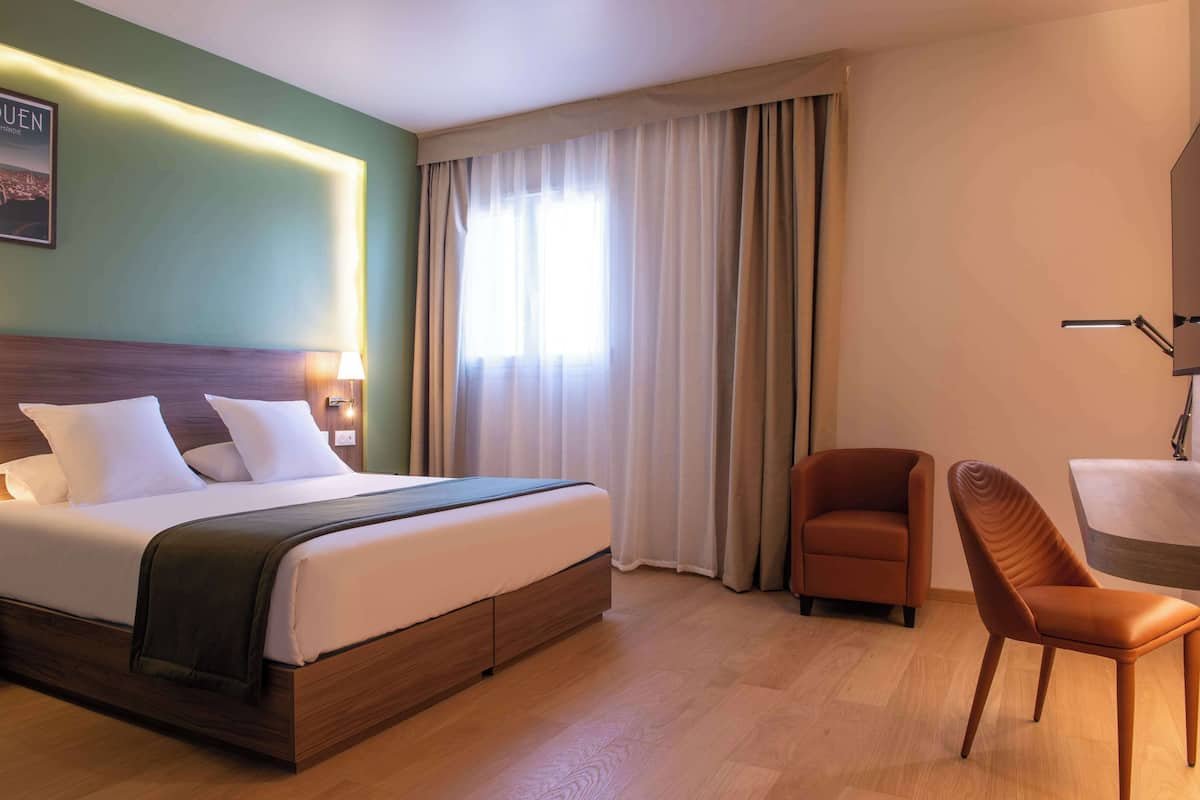 A hotel room with a large bed, a desk, two chairs, a lamp, and a window with curtains.