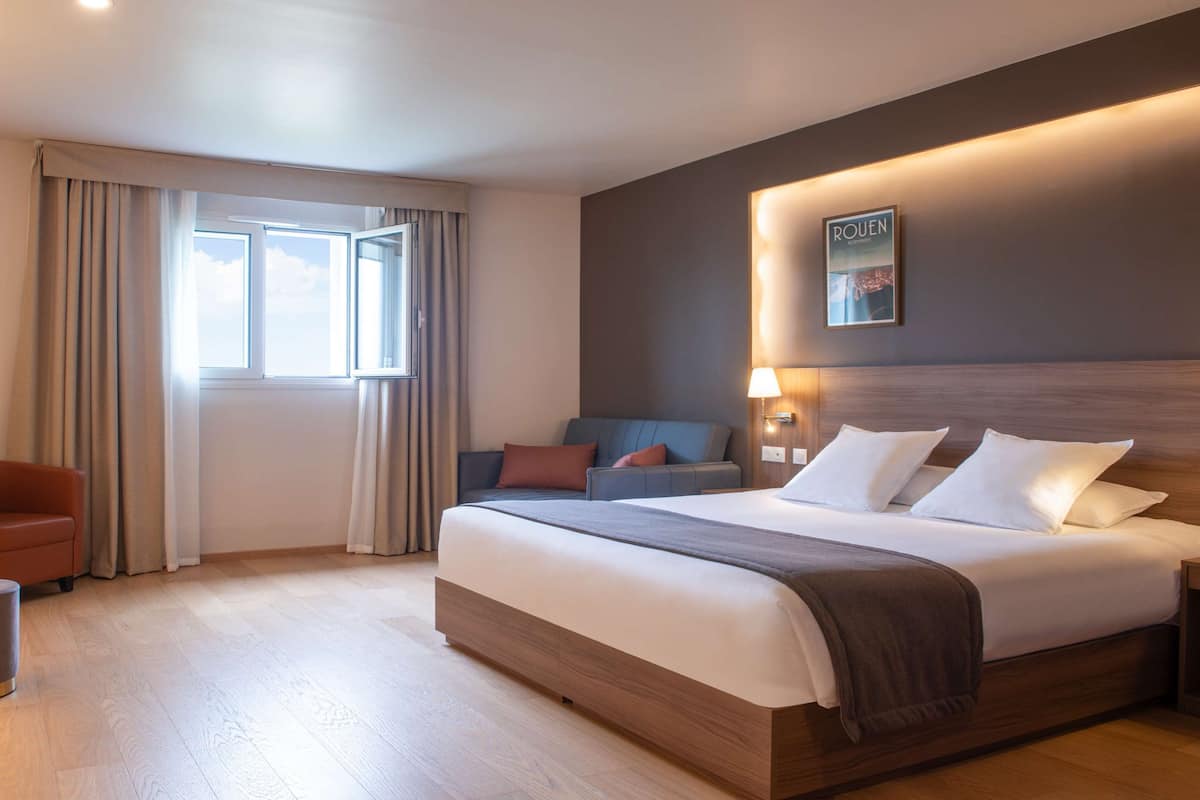 A hotel room with a bed, a seating area with a chair, a small table, a lamp, and a window with curtains.