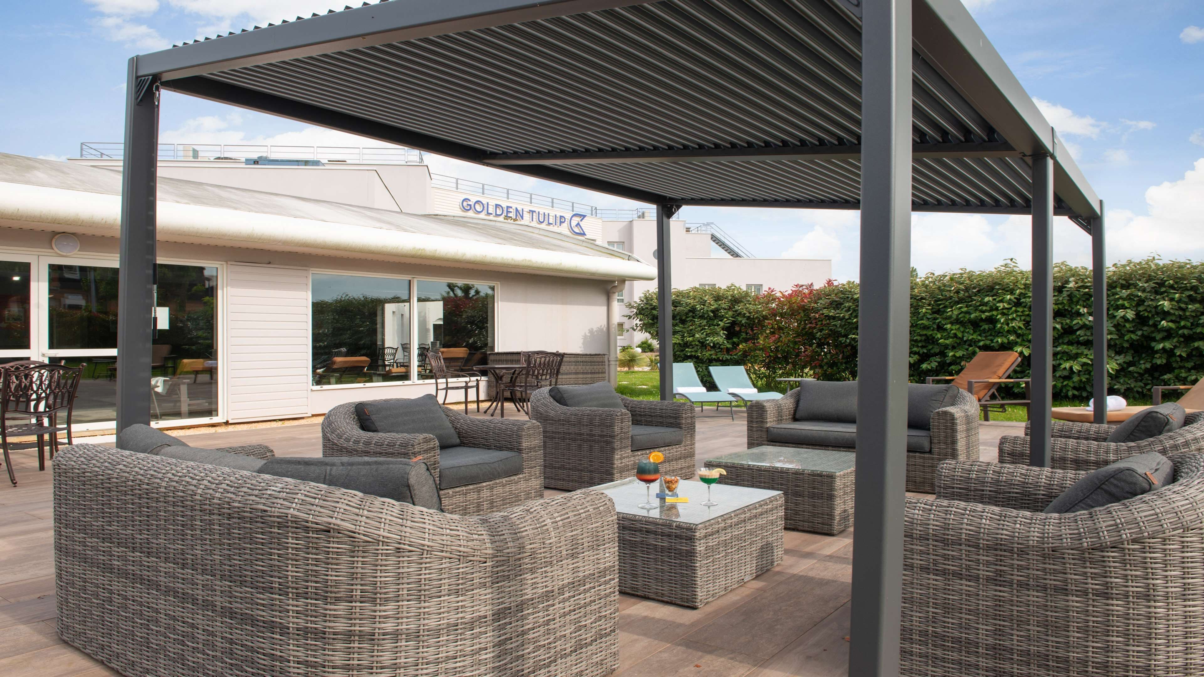 Un espace extérieur aménagé en terrasse, avec des meubles en osier, un coin salon abrité et, au fond, un bâtiment portant l’inscription « Golden Tulip ».