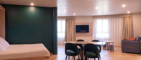 A modern hotel room with a bed, a dining area with chairs, a sofa, and a TV mounted on the wall.