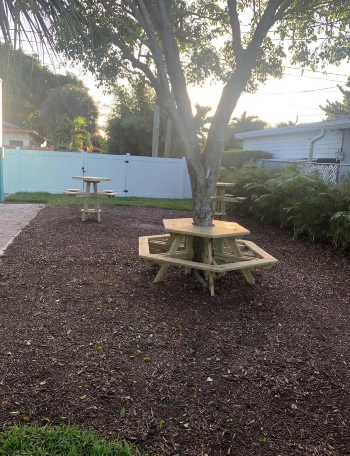 Una mesa de picnic rodeada de mantillo, un árbol y una cerca blanca en un patio trasero.