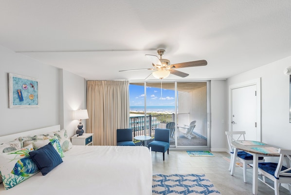 Habitación de hotel con cama, ventilador de techo, balcón con sillas y vista a la playa.