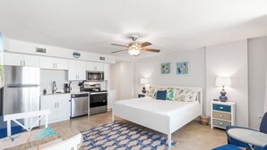 Une chambre d’hôtel compacte comprenant un coin cuisine, un lit avec une tête de lit à motifs, un ventilateur de plafond et une petite table avec une chaise.