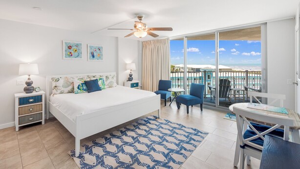 Une chambre avec un lit, des tables de chevet, un ventilateur de plafond, un balcon avec des chaises et une table, et une vue sur la plage.