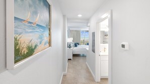 Un couloir mène à une chambre où se trouvent un lit, une table de chevet et un tableau représentant des voiliers accroché au mur.