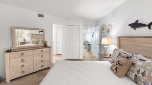 A bedroom with a wooden dresser, a bed with patterned pillows, a large mirror, and a wall-mounted fish decoration. - Sandcastle Condominiums Unit 513 (Port Aransas)