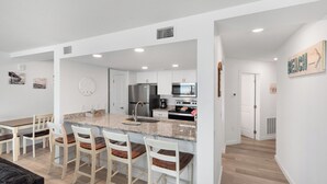 A modern kitchen with white cabinetry, a granite countertop, and stainless steel appliances. There are four bar stools with wooden seats and white backs. - Sandcastle Condominiums Unit 513 (Port Aransas)