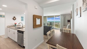 A modern kitchen with white cabinets, a dishwasher, and a granite countertop. There is a dining area with a wooden table and chairs, and a view of the ocean through large windows. - Sandcastle Condominiums Unit 513 (Port Aransas)