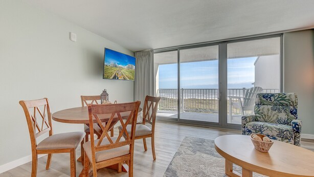 A spacious room with a wooden dining table and chairs, a patterned armchair, and a large window offering a view of the beach. - Sandcastle Condominiums Unit 523 (Port Aransas)