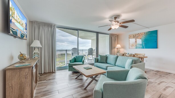 Een ruime woonkamer met een grote bank, een salontafel en een aan de muur bevestigde televisie. Via de glazen deuren is een balkon met buitenmeubilair zichtbaar.